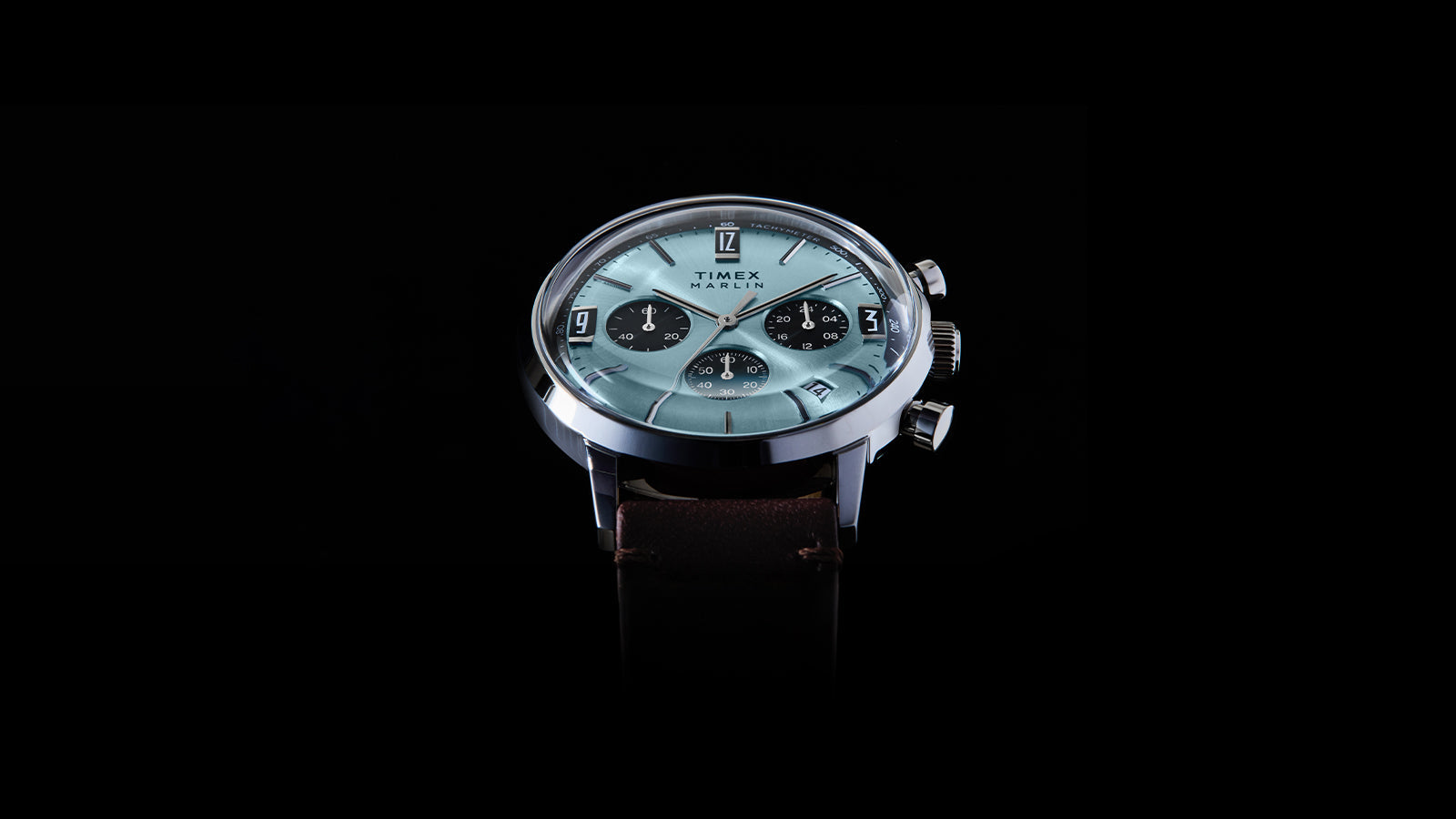 Angled view of the Timex Marlin Chronograph Tachymeter with a light blue dial, black subdials, and brown leather strap against a black background.