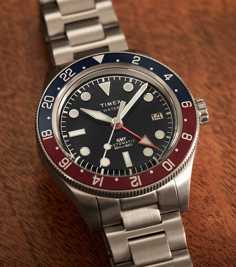 Timex Waterbury GMT automatic watch with stainless steel bracelet and black dial placed on a wooden surface.