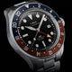 Close-up of Timex Waterbury GMT automatic watch featuring a black dial and red-blue bezel.