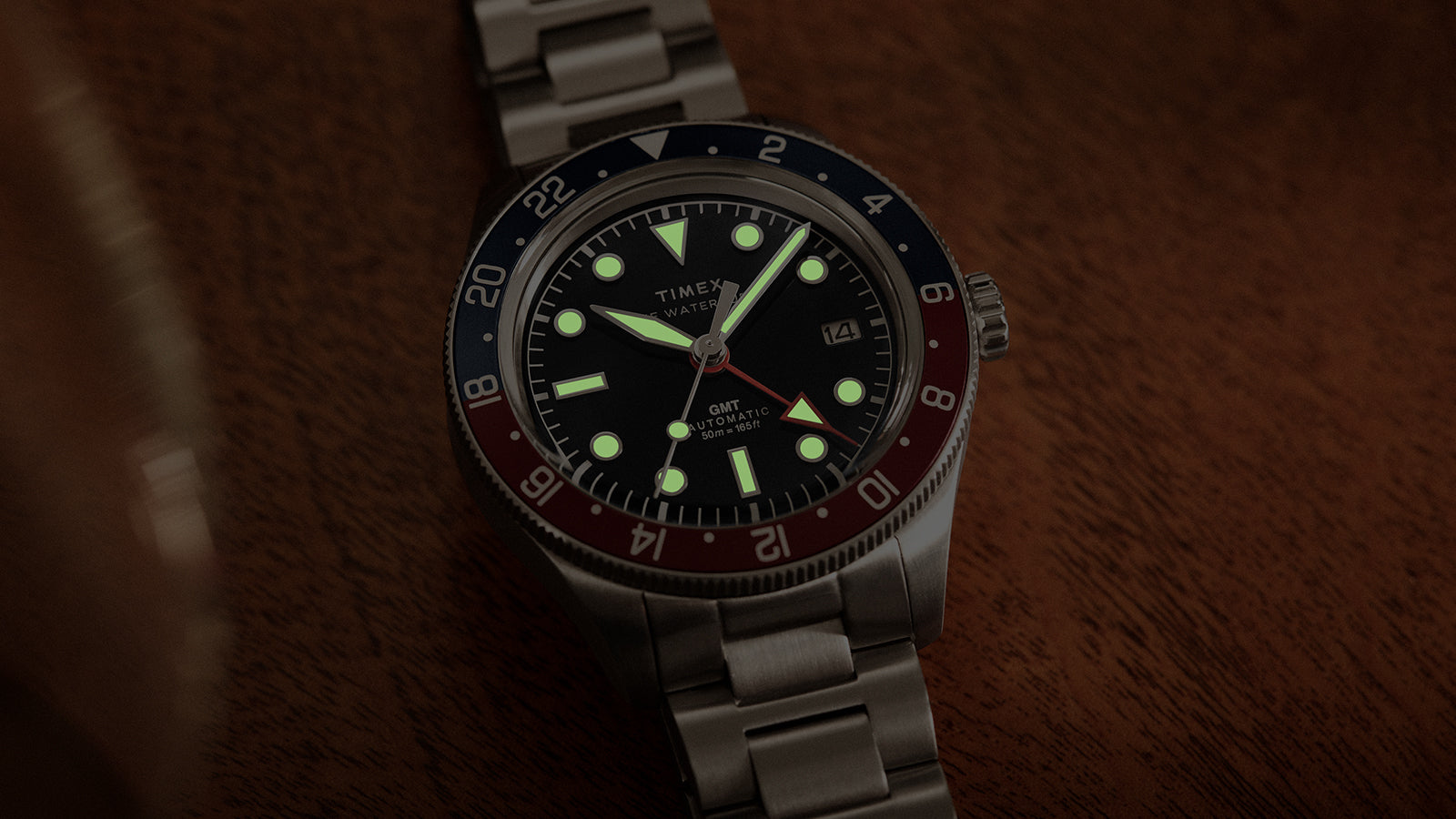 Timex Waterbury GMT automatic watch showing glowing luminous markers and hands in low light.