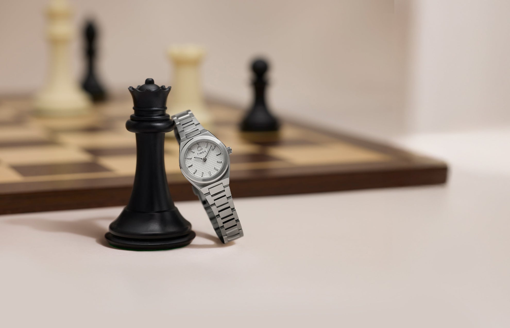 Q Timex® Continental Mini silver-tone watch with textured silver dial and stainless steel bracelet leaning against a black king chess piece with chessboard in background.
