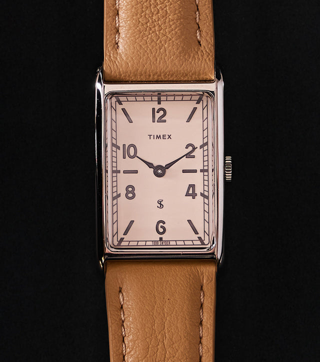 Timex x Todd Snyder 1976 Lexington rectangular watch with a cream dial and tan leather strap displayed vertically on a black background.