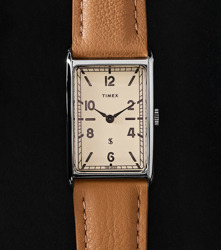 Timex x Todd Snyder 1976 Lexington rectangular watch with a cream dial and tan leather strap displayed vertically on a black background.