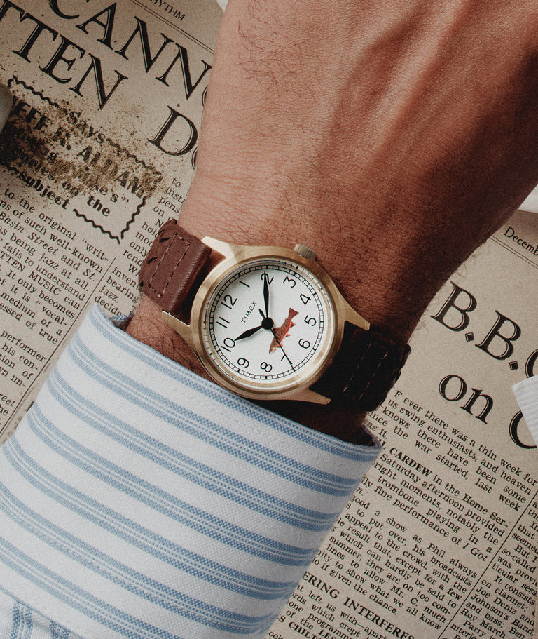 Timex MK1 for J.Crew on wrist with brown leather strap and cream dial, styled over vintage newspaper with blue striped shirt cuff. Brass-tone case and minimalist military-inspired design in warm natural light.