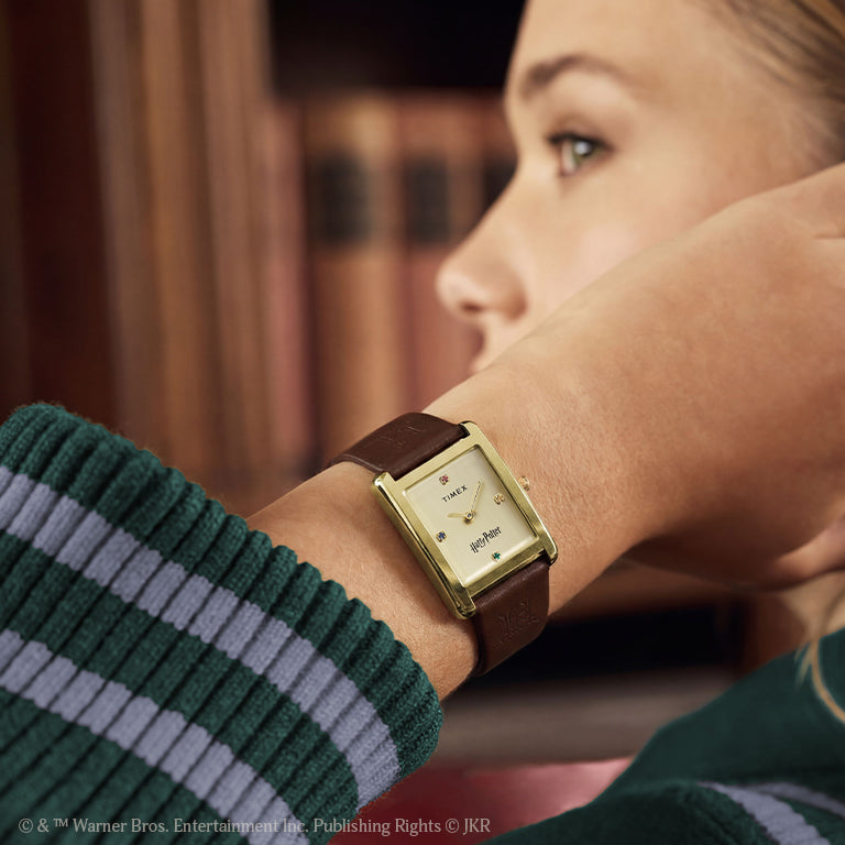 Close-up of a person wearing a gold-tone rectangular Timex watch with a brown leather strap, featuring a Harry Potter–themed dial, indoors with a softly blurred background.