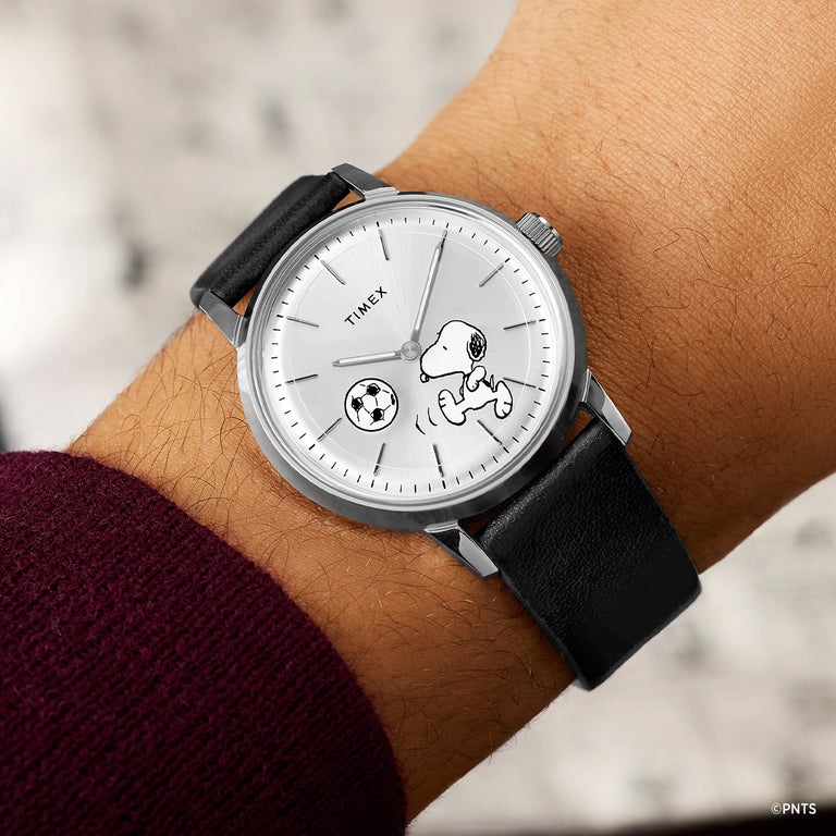 Peanuts® x Timex Marlin® Automatic Soccer watch with a silver dial, Snoopy soccer graphic, stainless-steel case, and black leather strap shown on wrist.