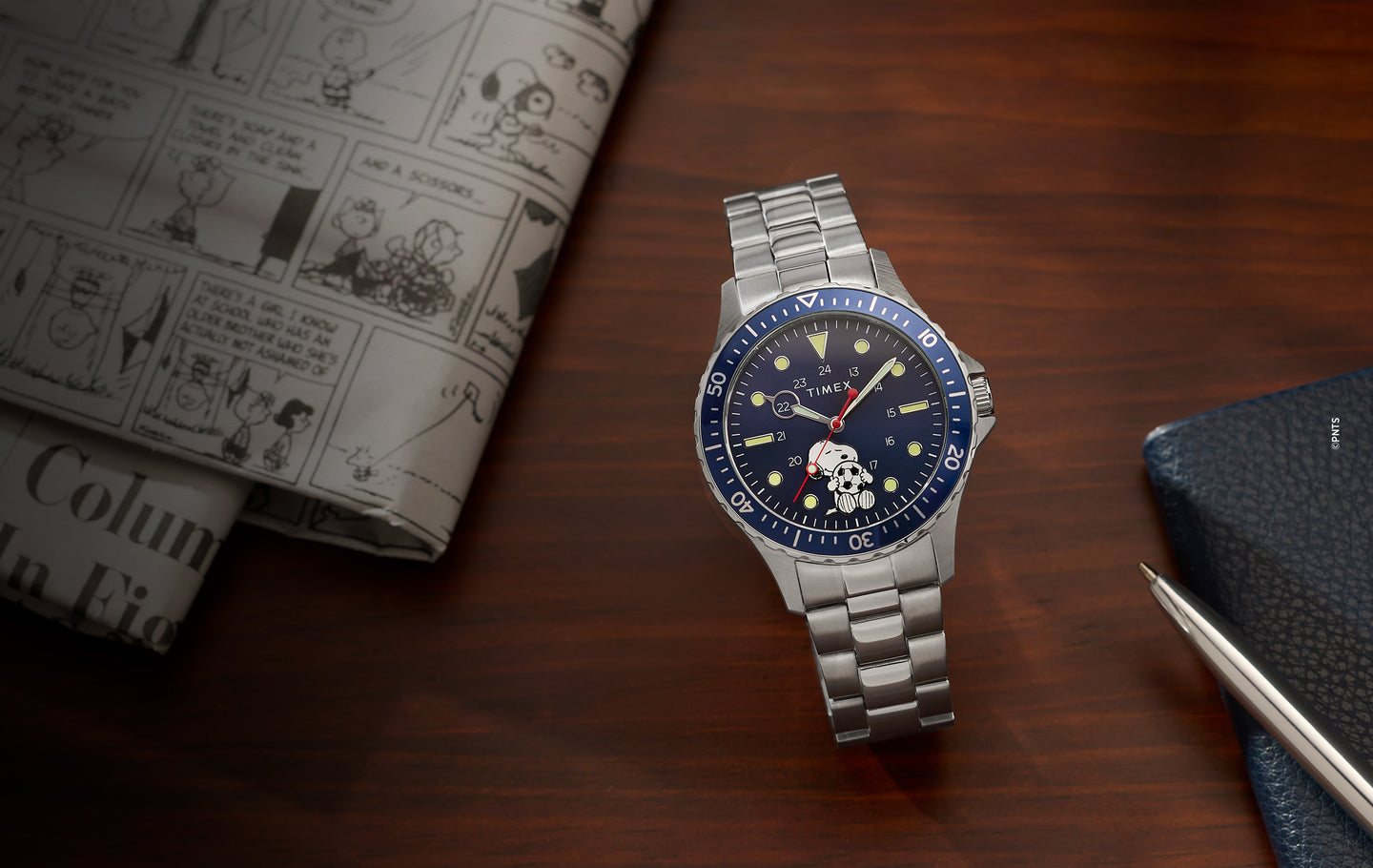 Peanuts x Timex Navi watch with Snoopy soccer dial on a stainless steel bracelet, placed on a wooden desk beside a Peanuts comic strip newspaper and notebook
