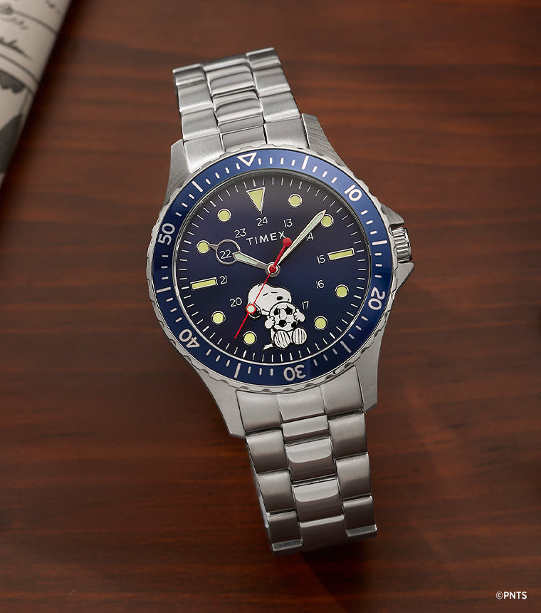 Peanuts x Timex Navi watch with Snoopy soccer dial on a stainless steel bracelet, placed on a wooden desk beside a Peanuts comic strip newspaper and notebook