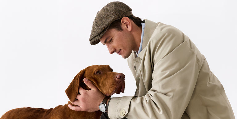 Man in beige trench coat and tweed flat cap holding a brown dog while wearing a Timex watch with brown leather strap against a clean studio background.