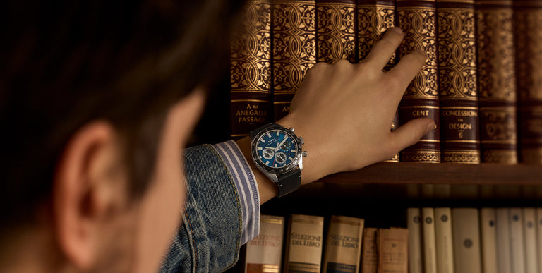 Person wearing a Timex Waterbury Traditional Chronograph watch with a blue dial and black leather strap, reaching toward a shelf of decorative books.