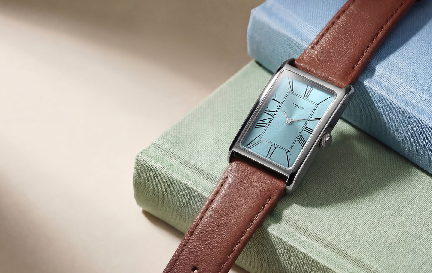 Timex® 1976 Lexington Reissue watch with a light blue rectangular dial, silver-tone case, and brown leather strap resting on pastel fabric blocks.