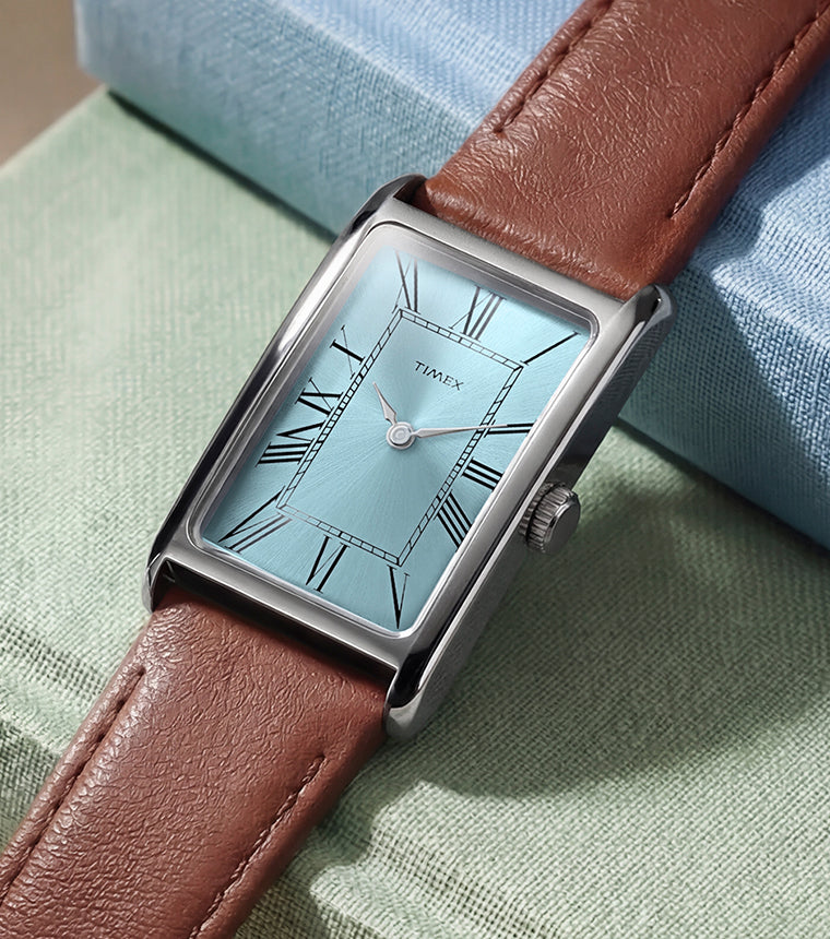 Timex® 1976 Lexington Reissue watch with a light blue rectangular dial, silver-tone case, and brown leather strap resting on pastel fabric blocks.