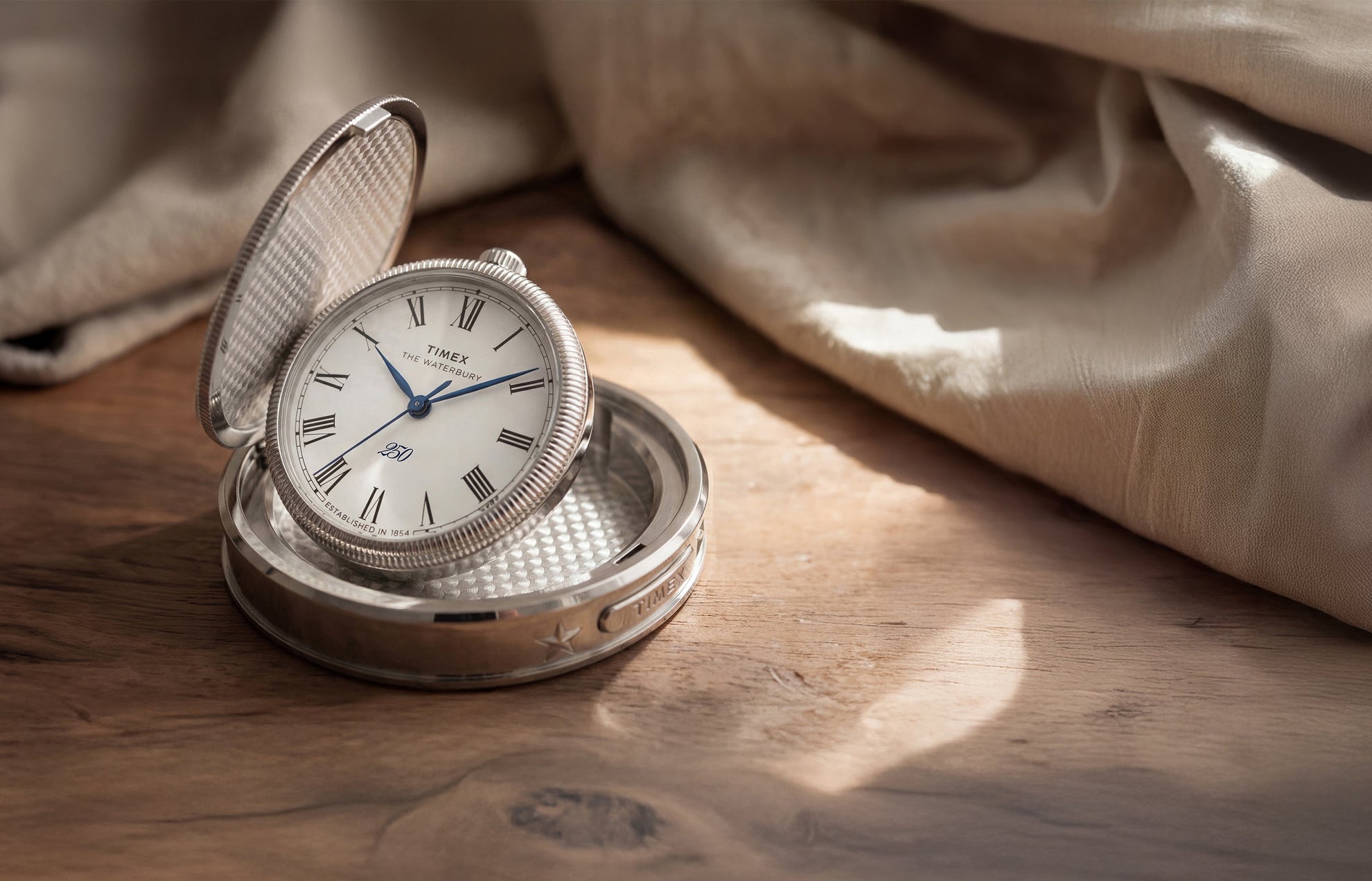 Waterbury America 250 Table Clock displayed open on a wooden table, featuring a silver-tone case, Roman numeral dial, and blue hands.