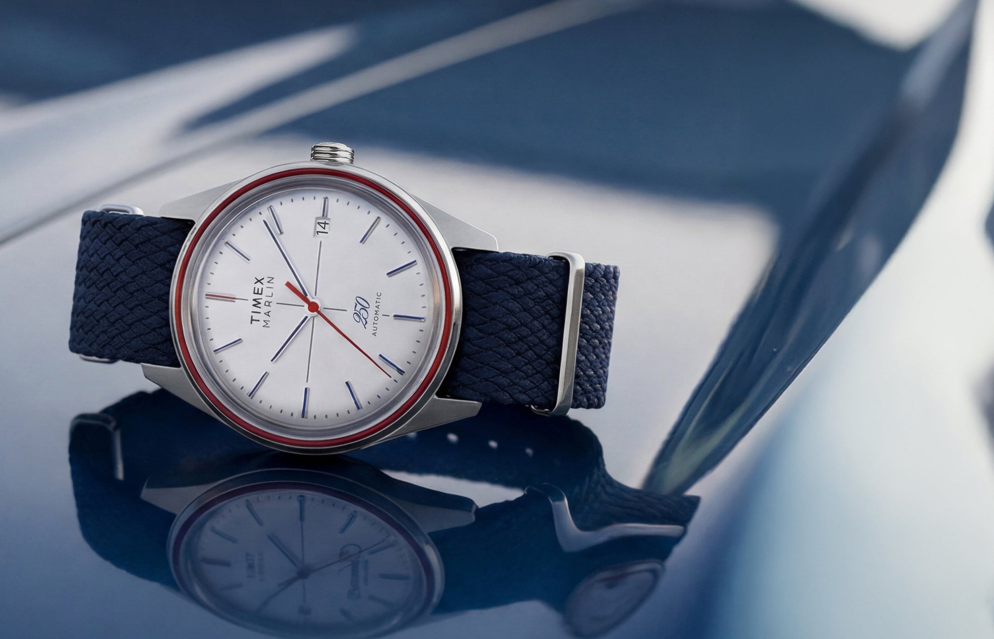 Marlin Jet America 250 automatic watch with a white dial, red bezel accent, red seconds hand, and blue woven strap displayed on a reflective blue surface.