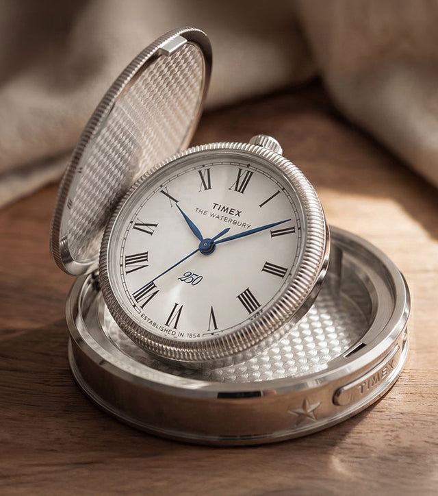 Waterbury America 250 Table Clock displayed open on a wooden table, featuring a silver-tone case, Roman numeral dial, and blue hands.