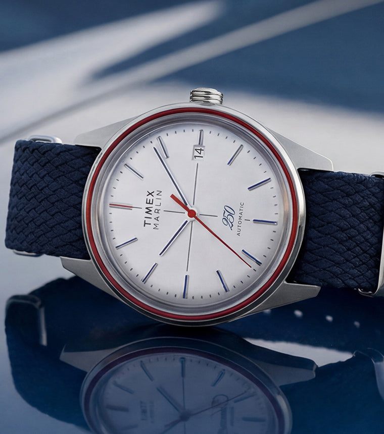 Marlin Jet America 250 automatic watch with a white dial, red bezel accent, red seconds hand, and blue woven strap displayed on a reflective blue surface.