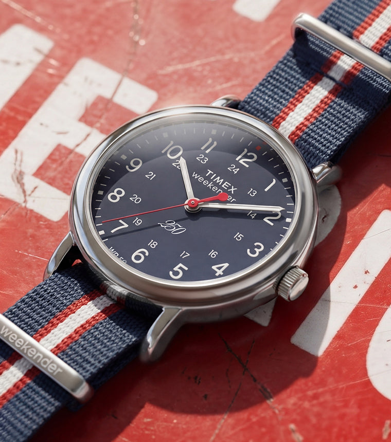 Weekender America 250 watch with a blue dial, red seconds hand, and red, white, and blue striped fabric strap displayed on a vintage red American-themed surface.