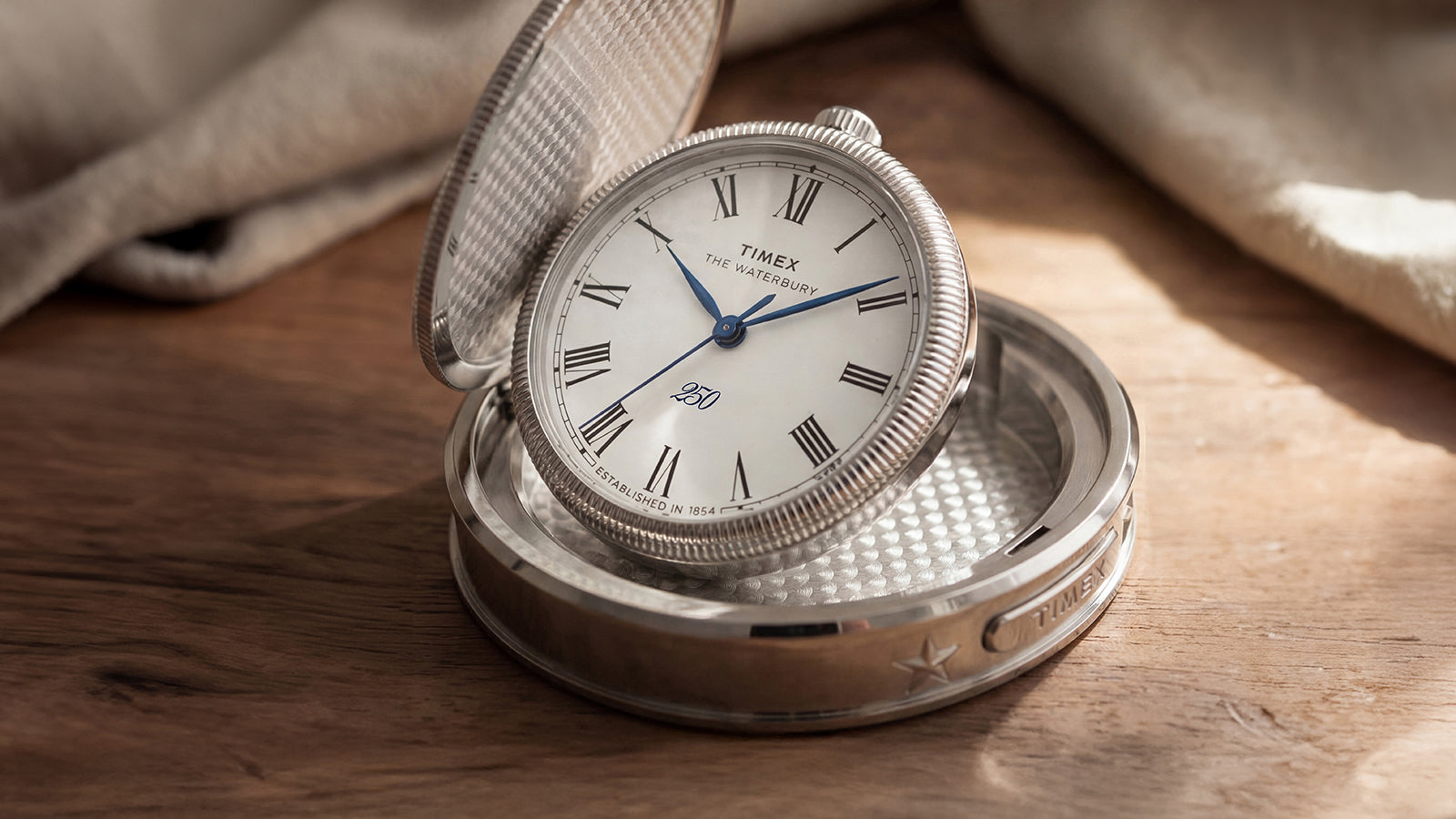 Waterbury America 250 Table Clock open to reveal a white Roman numeral dial with blue hands and a silver-tone hinged case.