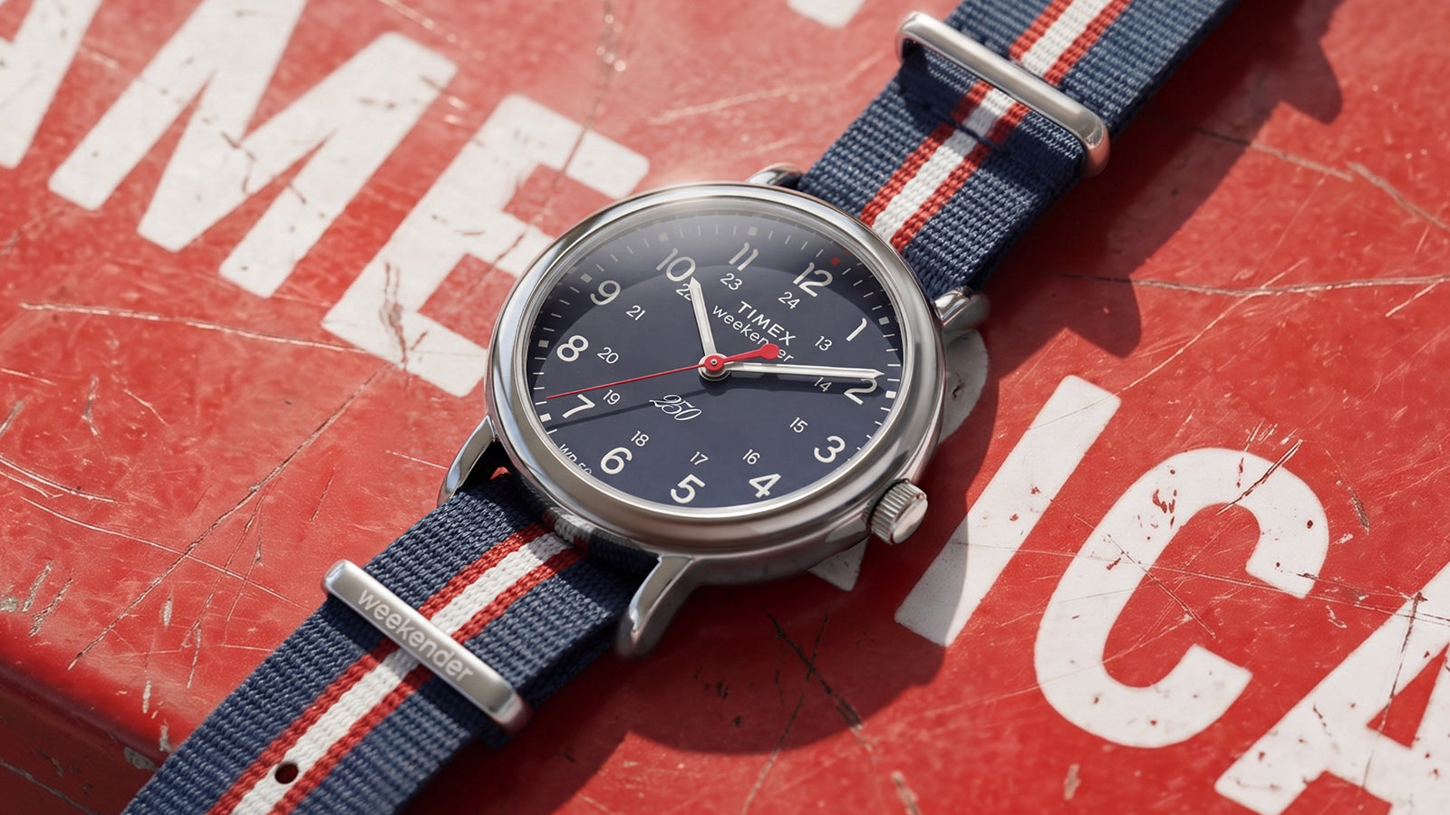 Weekender America 250 watch featuring a blue dial, white Arabic numerals, red seconds hand, and patriotic striped fabric strap.