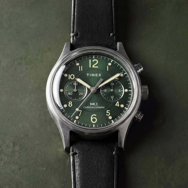 Timex MK1® Chronograph watch with a green dial, brushed gray case, black leather strap, and dual chronograph sub-dials, displayed on a green background.