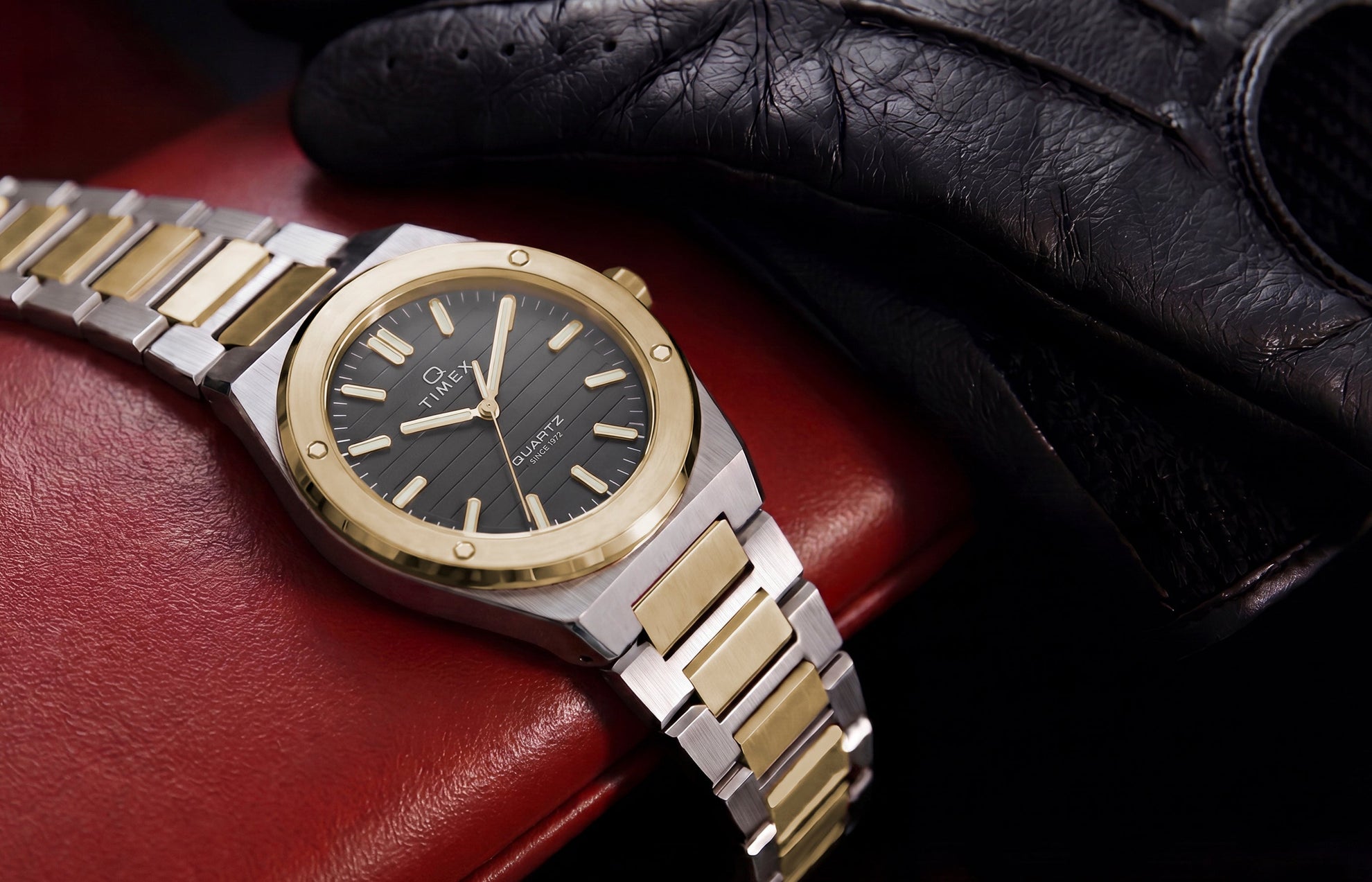 Timex Q Marbella two-tone stainless steel watch with a black textured dial displayed on a red leather car seat beside a black driving glove.