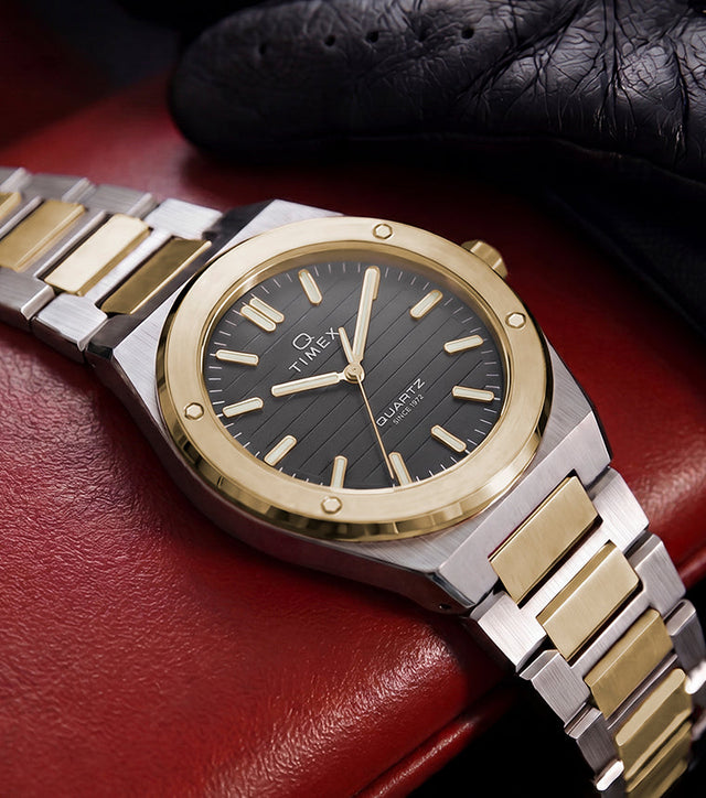 Timex Q Marbella two-tone stainless steel watch with a black textured dial displayed on a red leather car seat beside a black driving glove.