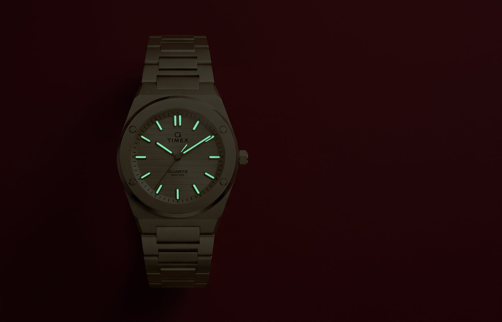 Q Timex Marbella gold-tone stainless steel watch with glowing Indiglo® hands and hour markers against a red background.