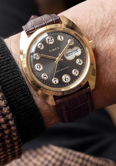 Timex® 1976 Rotary Phone Dial Reissue with a gold-tone case, black dial, and brown leather strap worn on the wrist with a patterned blazer.