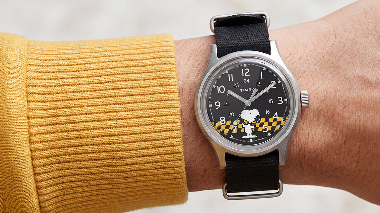 Peanuts® x Timex MK1® watch worn with a black fabric strap, featuring a black dial with Snoopy in sunglasses and yellow racing-inspired accents.