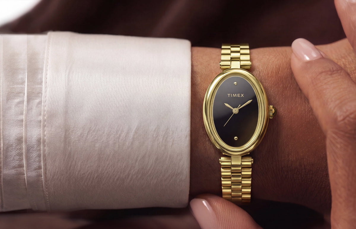 TW2Y86200 Person wearing the Timex Cavatina® Luxe gold-tone watch with a black dial while seated outdoors near the water, styled with a white outfit and sunglasses.