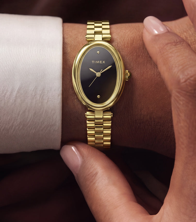 TW2Y86200 Person wearing the Timex Cavatina® Luxe gold-tone watch with a black dial while seated outdoors near the water, styled with a white outfit and sunglasses.