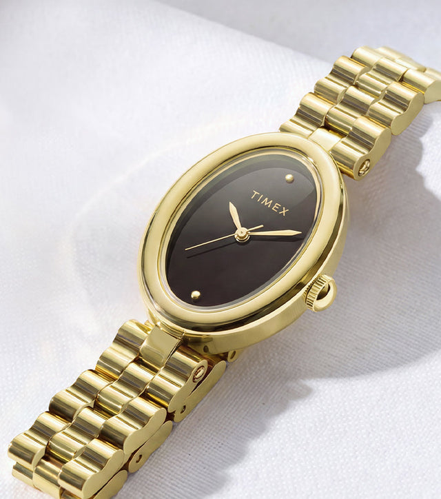 Timex Cavatina® Luxe watch with a gold-tone stainless steel bracelet, oval case, and black dial displayed on soft white fabric.