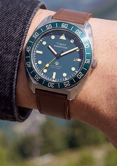 Timex Expedition Pioneer Automatic worn on the wrist with a dark jacket, featuring a blue dial, blue rotating bezel, and brown leather strap.