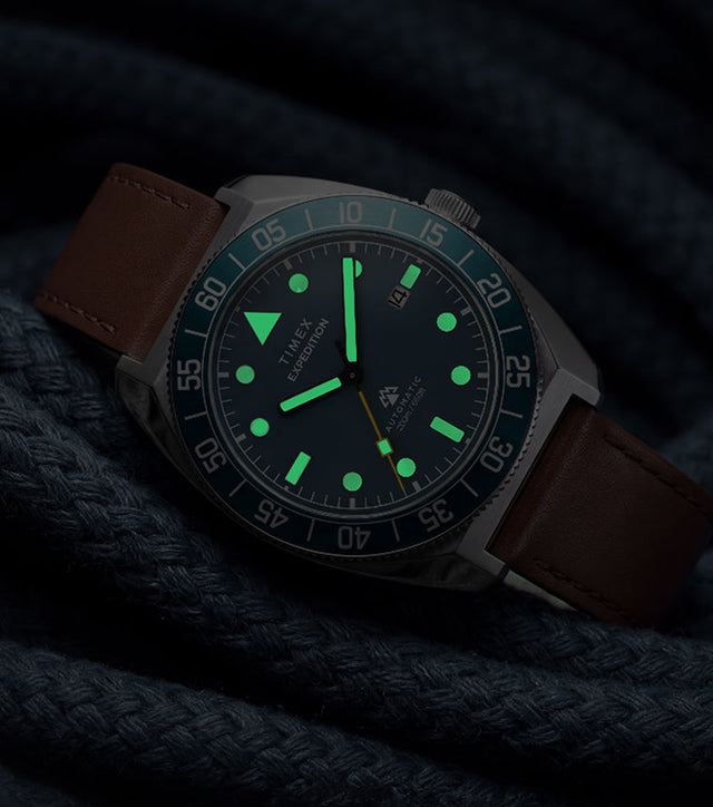 Timex Expedition Pioneer Automatic in low light with green luminous hands and hour markers glowing against coiled navy rope.