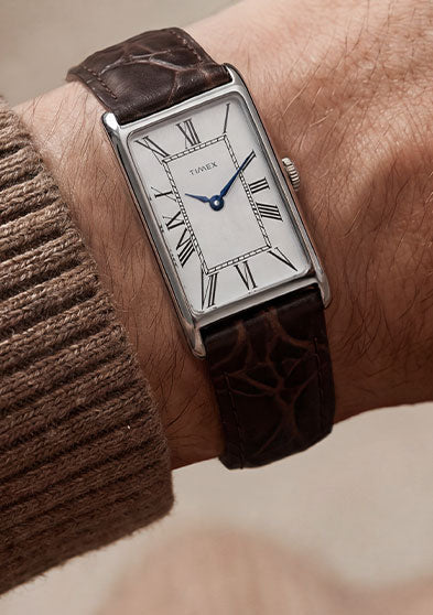 Timex 1976 Lexington Reissue Inspired watch worn on the wrist, featuring a silver sunray dial with Roman numerals, blue hands, and a brown crocodile-grain leather strap.