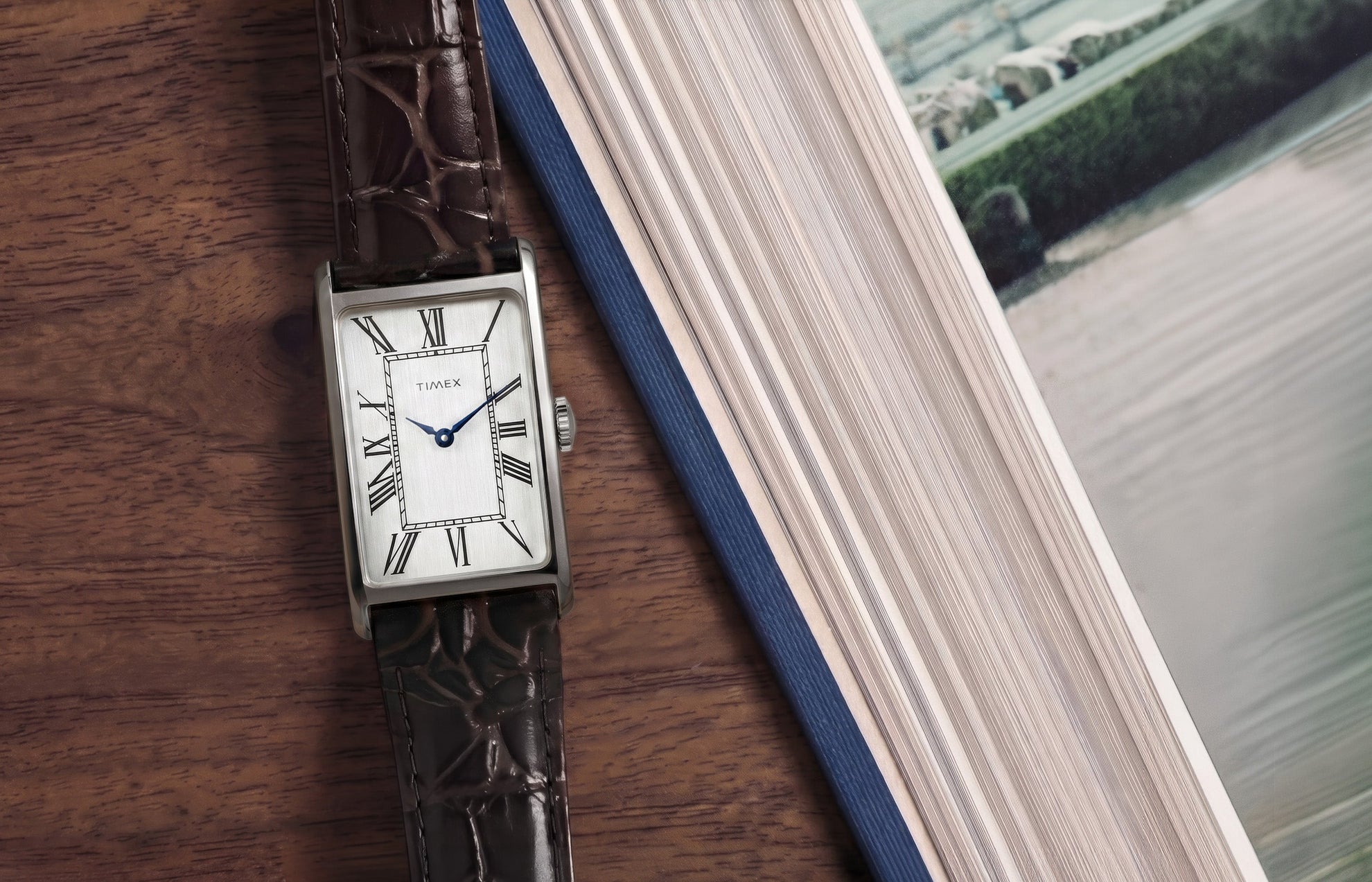 Timex 1976 Lexington Reissue Inspired watch with a rectangular silver-tone case, silver dial, blue hands, and brown crocodile-grain leather strap resting beside an open book on a wooden table.