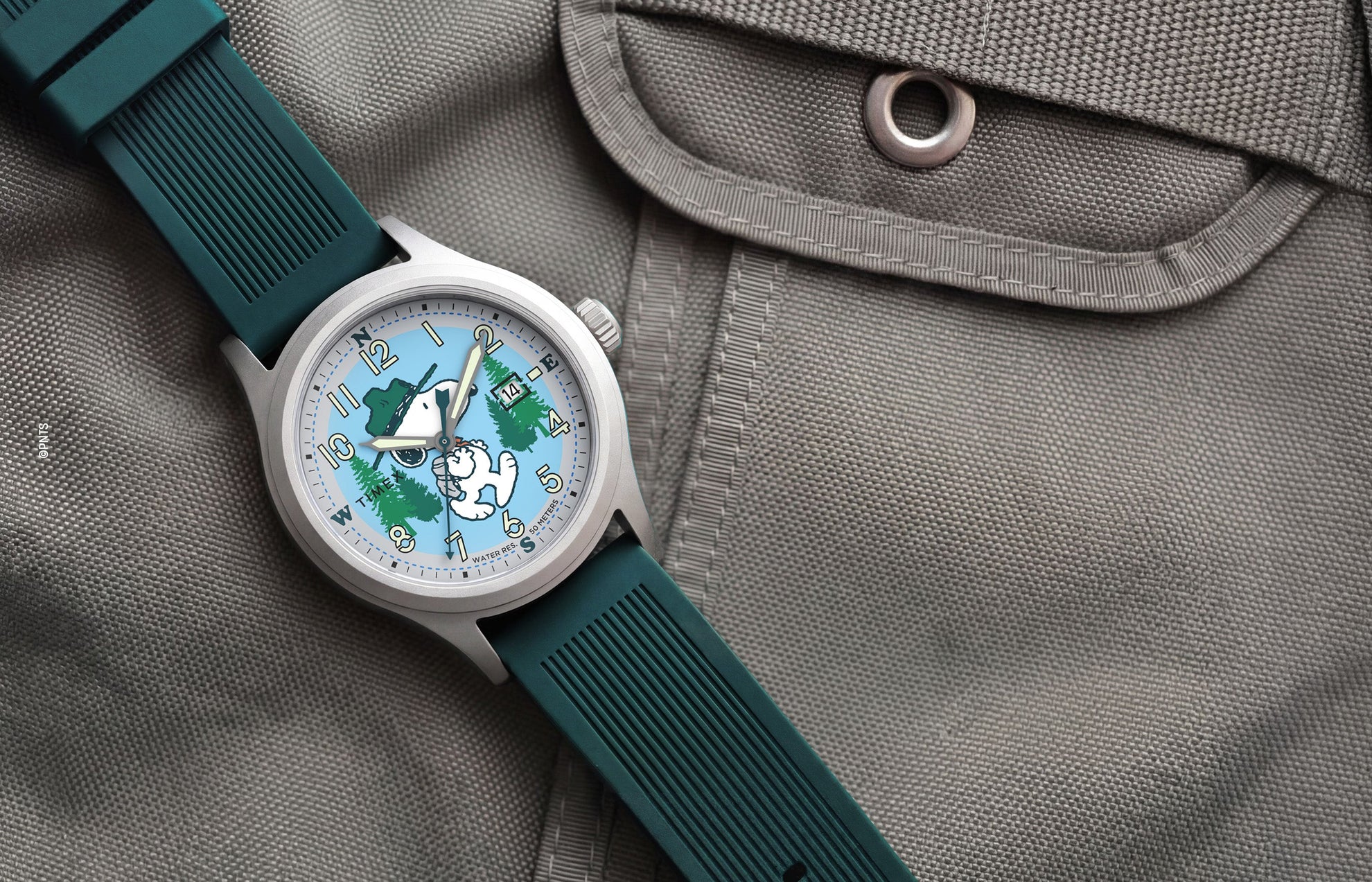Peanuts® x Timex Expedition® Scout watch with a silver-tone case, light blue dial, and dark green silicone strap, featuring Snoopy hiking through a forest scene, luminous Arabic numerals, a date window, and displayed on a gray tactical fabric background.