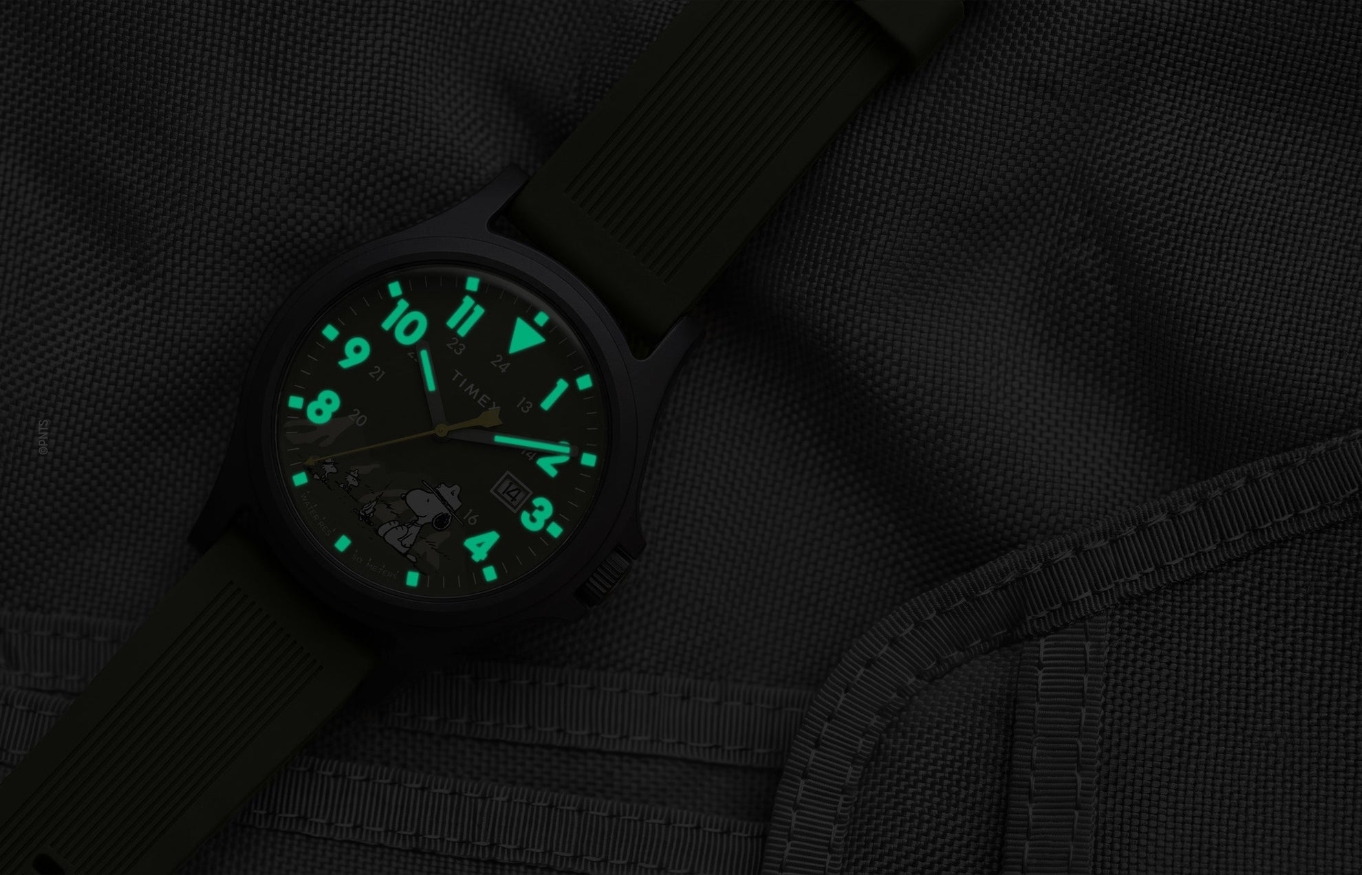 Peanuts® x Timex Expedition® Acadia watch with illuminated green Indiglo® dial, highlighting the glowing Arabic numerals, hour markers, and hands against a dark gray tactical fabric background.