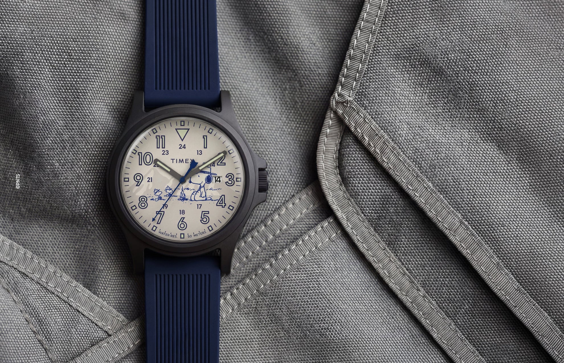 Peanuts® x Timex Expedition® Acadia watch with a matte black case, cream dial, and navy blue silicone strap, featuring Snoopy with Woodstock artwork, bold blue Arabic numerals, a date window, and blue hands displayed on a gray tactical fabric background.