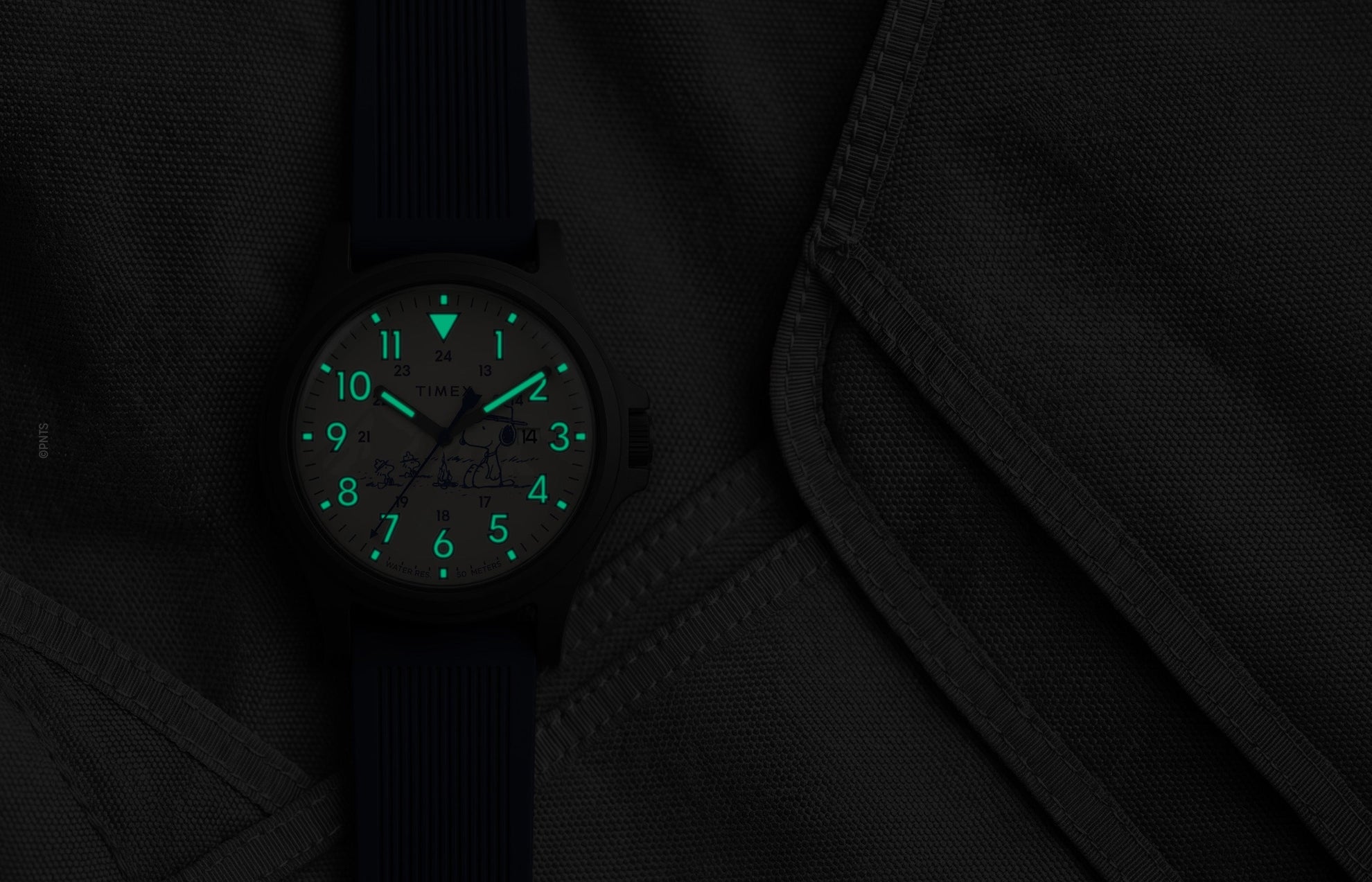 Peanuts® x Timex Expedition® Acadia watch with illuminated green Indiglo® dial, highlighting the glowing Arabic numerals, hour markers, and hands against a dark gray tactical fabric background.