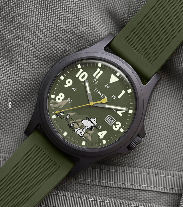 Peanuts® x Timex Expedition® Acadia watch with a matte black case, olive green dial and silicone strap, featuring Snoopy in camouflage artwork, bold Arabic numerals, a date window, and yellow seconds hand displayed on a gray tactical fabric background.