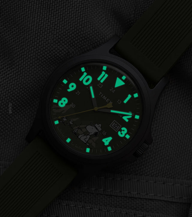 Peanuts® x Timex Expedition® Acadia watch with illuminated green Indiglo® dial, highlighting the glowing Arabic numerals, hour markers, and hands against a dark gray tactical fabric background.