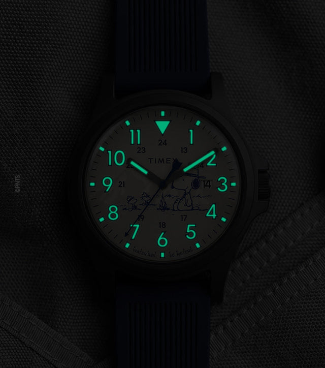 Peanuts® x Timex Expedition® Acadia watch with illuminated green Indiglo® dial, highlighting the glowing Arabic numerals, hour markers, and hands against a dark gray tactical fabric background.