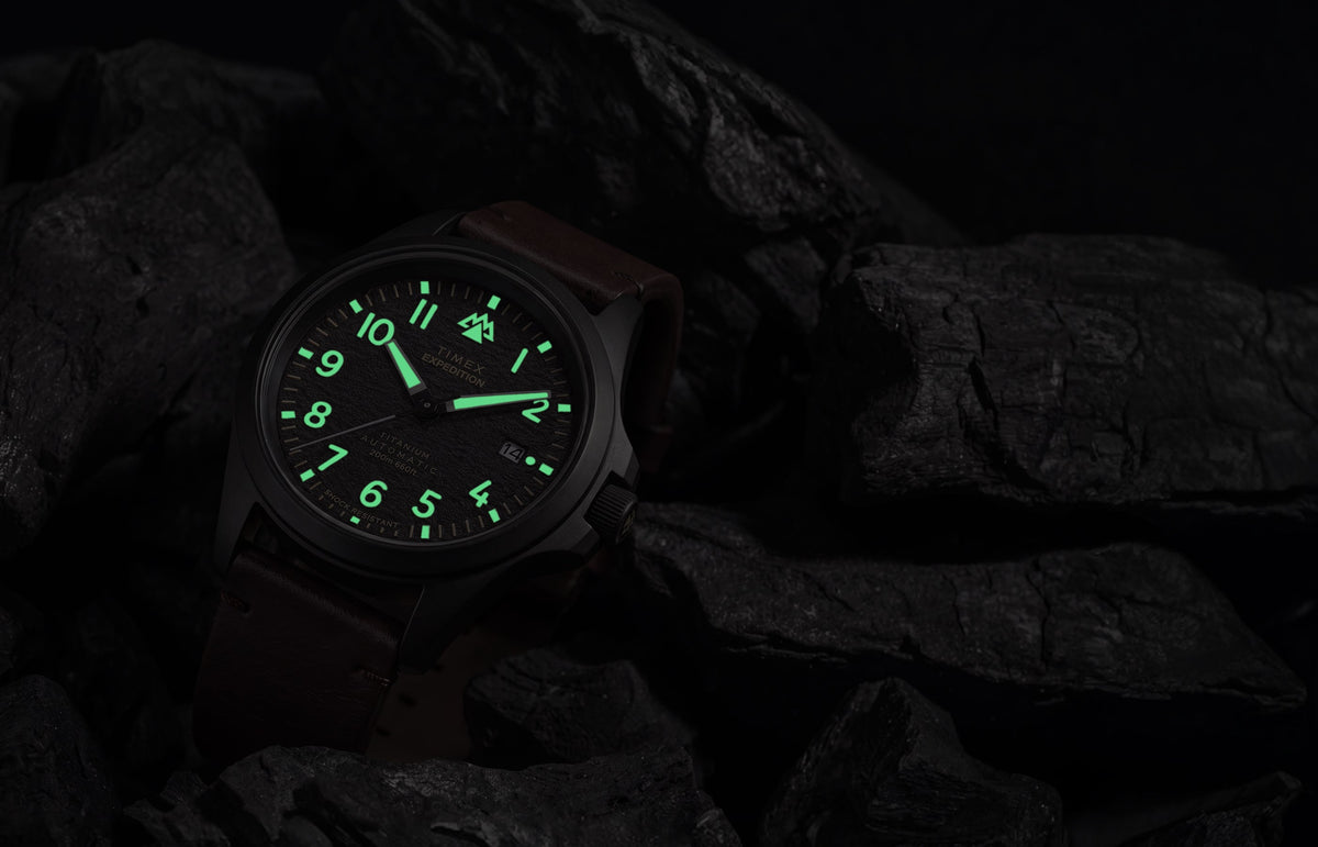 Expedition® Field Post Automatic titanium watch with glowing luminous hands and hour markers in low light.