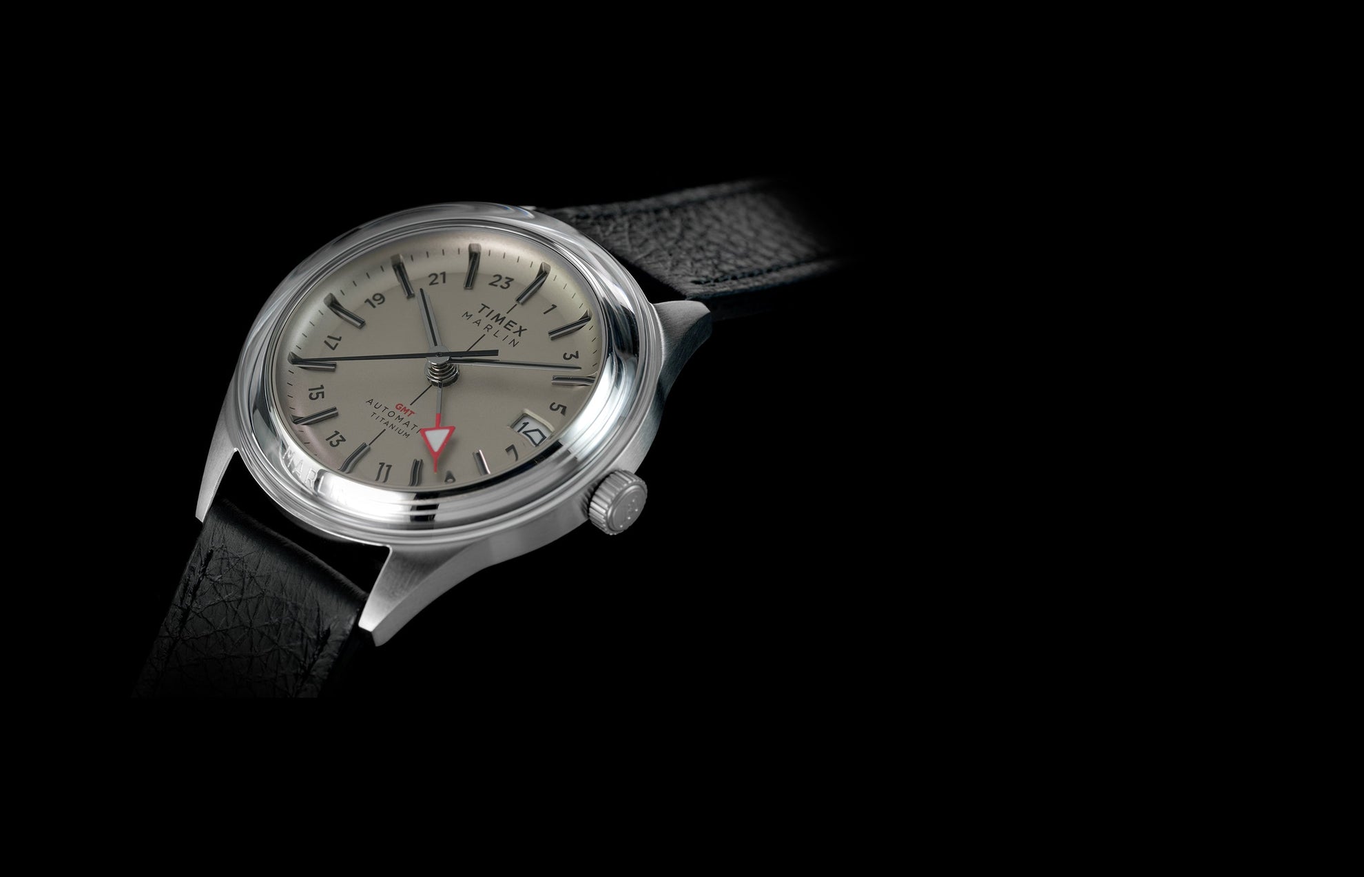 Timex Marlin® Jet Titanium Automatic GMT watch with silver-tone titanium case, black leather strap, cream dial, red GMT hand, and date window on a black background.