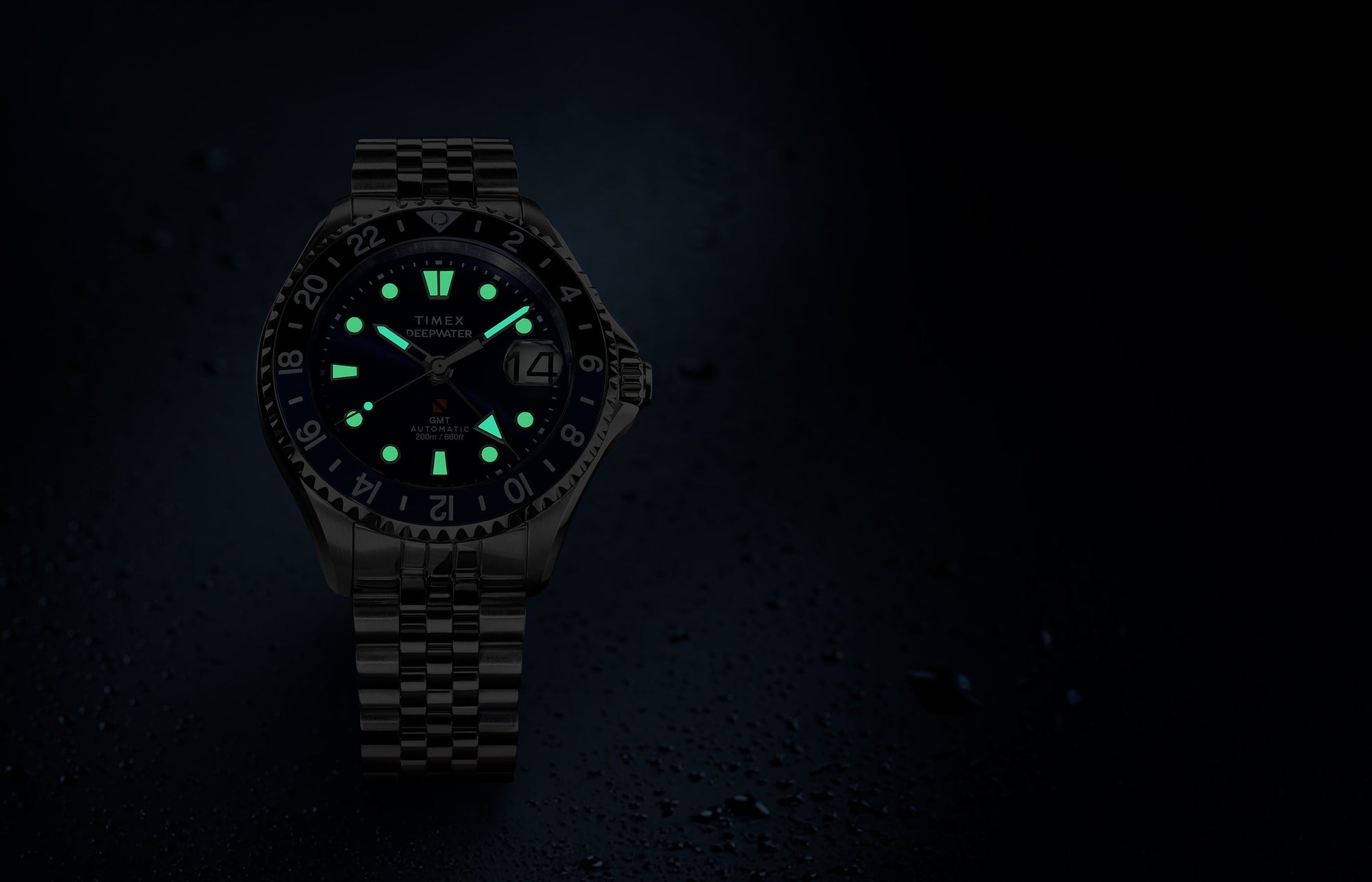 Timex Deepwater Meridian 200 Automatic GMT watch with a blue dial and glowing luminous hands and hour markers, displayed against a dark, water-droplet background.