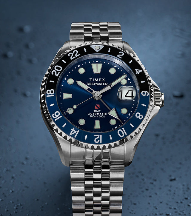 Timex Deepwater Meridian 200 Automatic GMT stainless steel watch featuring a blue sunray dial, GMT bezel, magnified date window, and jubilee-style bracelet on a textured blue background.