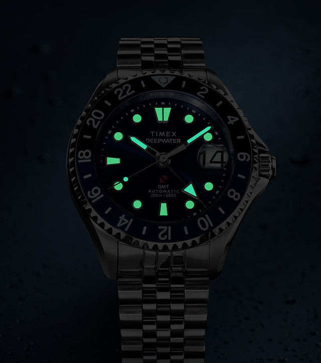 Timex Deepwater Meridian 200 Automatic GMT watch with a blue dial and glowing luminous hands and hour markers, displayed against a dark, water-droplet background.