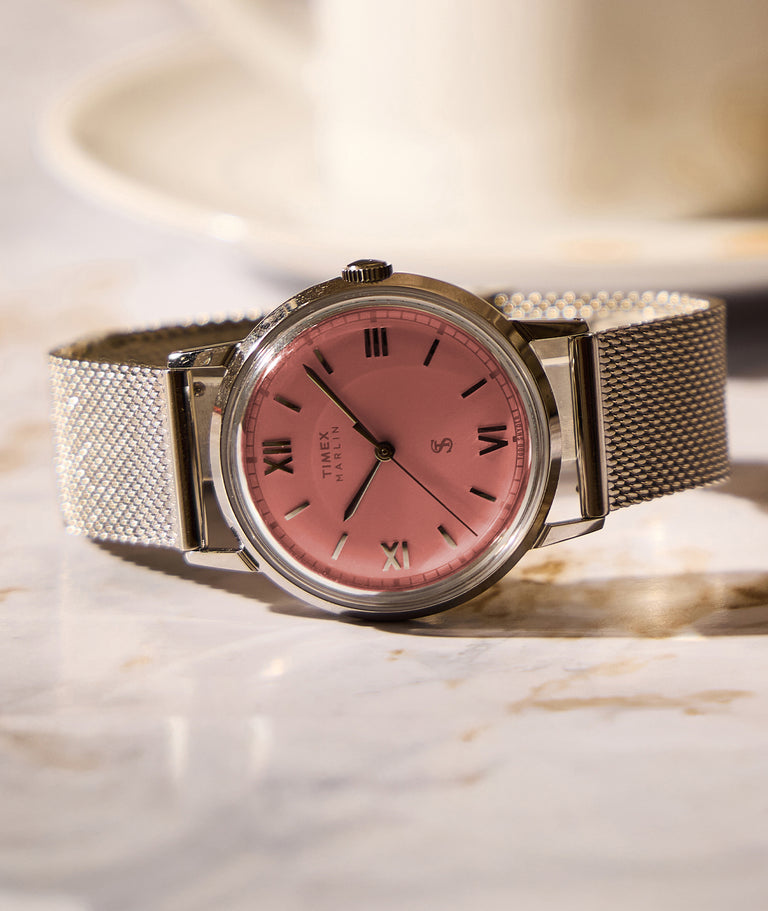 Todd Snyder x Timex Marlin® Mesh watch with a pink dial, silver-tone case, Roman numeral markers, and a silver mesh bracelet, resting on a marble surface.