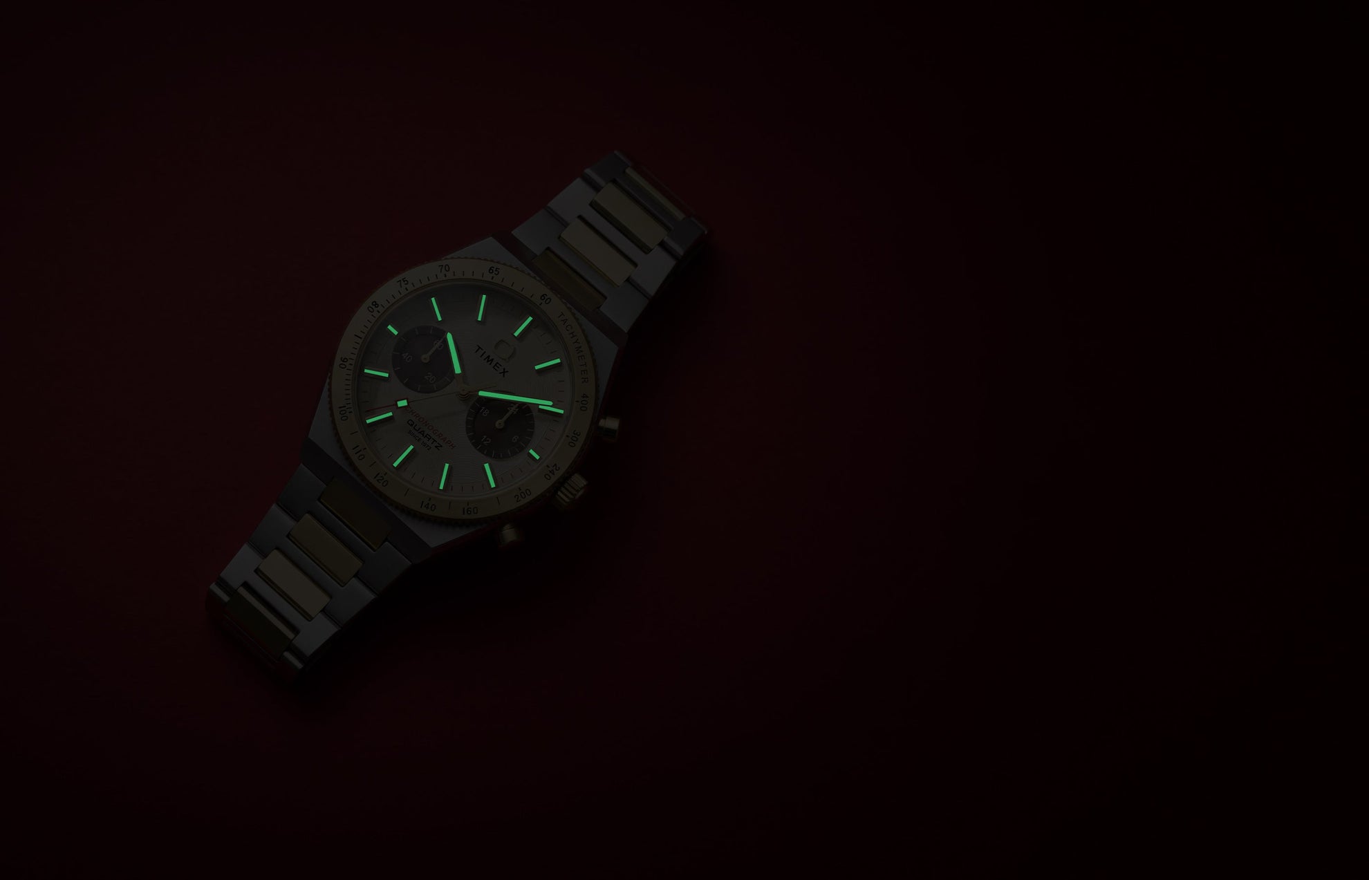 Q Timex Continental Chronograph on a dark red background with green luminous hands and hour markers visible in low light.